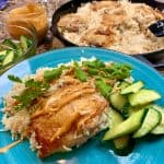 A blue plate with chicken and rice garnished with cilantro and sauce, accompanied by slices of cucumber. In the background, a pan filled with rice and chicken and a small bowl of sauce.