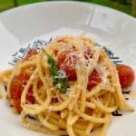 A plate of spaghetti topped with cherry tomatoes, shredded cheese, and a sprig of basil. The dish is served in a white bowl with a decorative black design, set outdoors against a blurred green background.