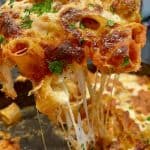 Close-up of a gooey, cheesy baked pasta dish. Rigatoni pasta is covered with melted cheese and sauce, with pieces of meat and garnished with parsley. A slice is being lifted, showcasing stretchy melted cheese.