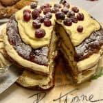 A round cake with creamy layers, topped with powdered sugar and cranberries, sits on a plate with the word Panettone written on it. A slice is cut out, revealing the moist interior. In the background, there are pastries and a striped candle.
