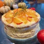 A layered cake topped with peach slices and a sprig of rosemary on a glass plate. The cake is surrounded by small pumpkins and red apples, giving an autumnal feel.