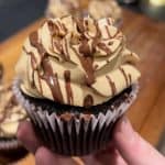 A hand holds a chocolate cupcake topped with swirls of light brown frosting and drizzled with chocolate. The background is blurred, showing more cupcakes on a wooden surface.