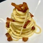 A plate of spaghetti carbonara is neatly presented in a swirl topped with crispy bacon pieces. The creamy pasta is lightly coated in sauce and garnished with black pepper, served on a white dish.