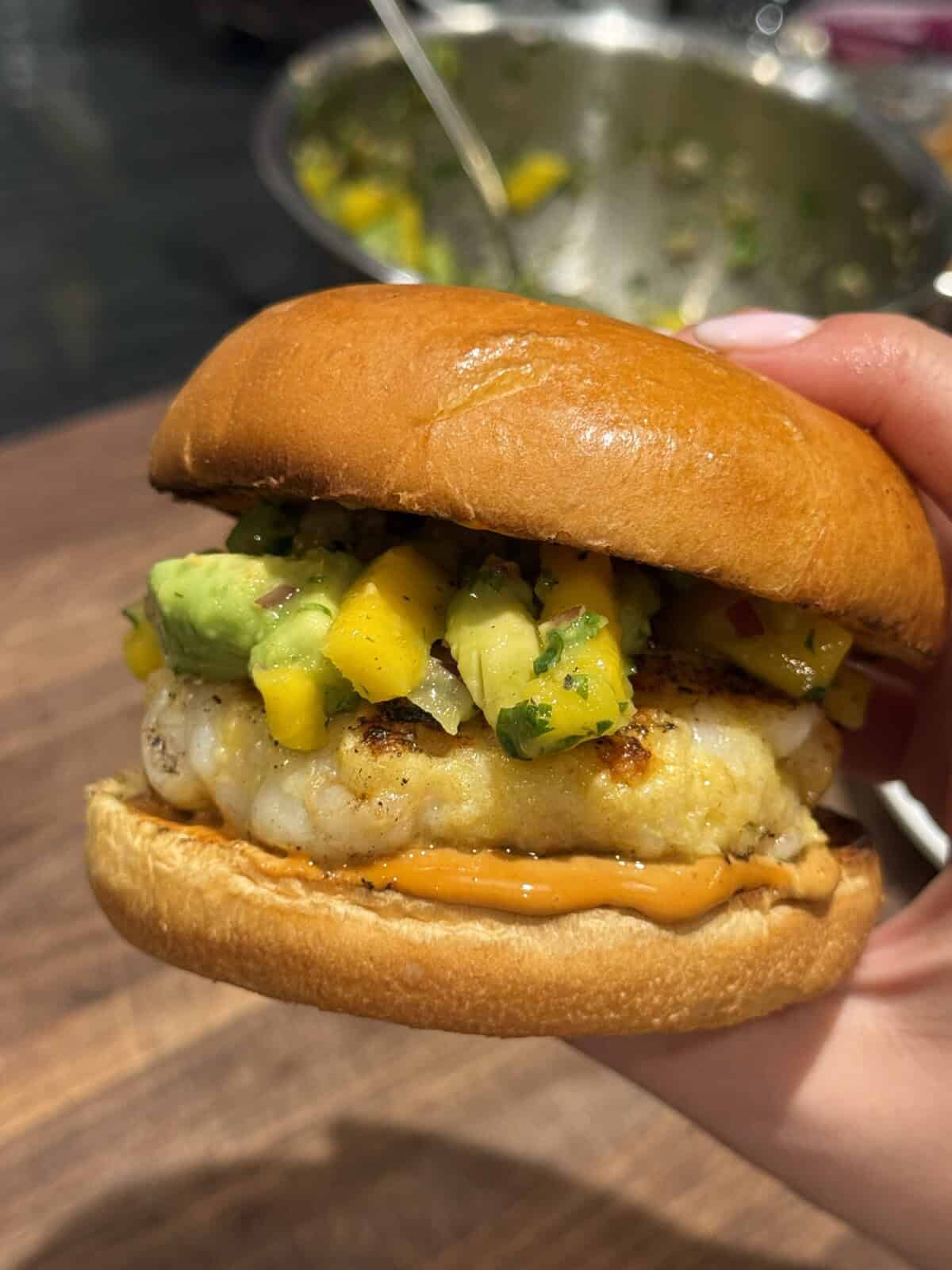 A hand holds a sandwich with a grilled patty, topped with a colorful mango and avocado salsa, and orange sauce on a soft, shiny bun. A bowl with more salsa is blurred in the background.