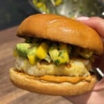 A hand holds a sandwich with a grilled patty, topped with a colorful mango and avocado salsa, and orange sauce on a soft, shiny bun. A bowl with more salsa is blurred in the background.