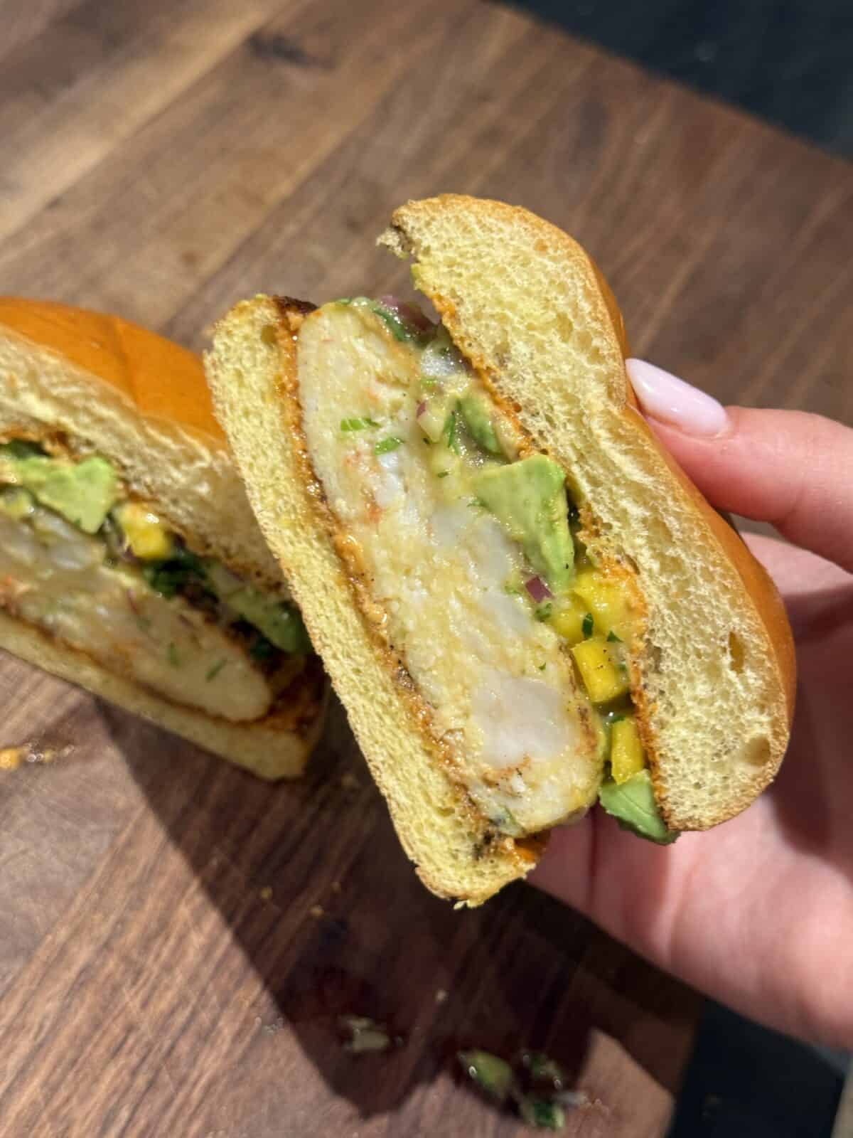 A hand holds half of a sandwich with a thick piece of breaded chicken, sliced avocado, chopped herbs, and a soft bun, shown on a wooden surface.