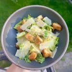 A bowl of Caesar salad with romaine lettuce, croutons, and shaved Parmesan cheese. The dish is held up outdoors with a green lawn in the background.