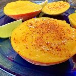 Slices of mango sprinkled with chili powder are on a blue plate, accompanied by lime wedges. The mangos vibrant orange contrasts with the blue dish, and the chili adds texture.