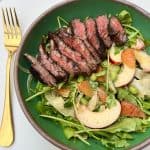 A green bowl filled with a fresh salad topped with slices of medium-rare steak. The salad includes mixed greens, peach slices, citrus segments, edamame, and a light dressing. A gold fork is placed on the white table beside the bowl.