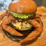A unique burger on a black plate with a whole deep-fried soft-shell crab protruding from the sides. It includes avocado slices, colorful slaw, and a glossy bun, all set on a wooden surface.