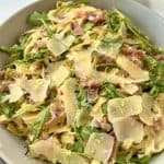 A bowl of tagliatelle pasta topped with arugula, thinly sliced prosciutto, and shaved Parmesan cheese. The dish is garnished with lemon zest, creating a fresh and colorful appearance. A cup is partially visible in the background.