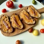 Grilled bread slices topped with shredded tomatoes and anchovies on a wooden board, surrounded by various small and large tomatoes on a round white table.