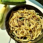 A pan of spaghetti with sliced zucchini and herbs, set on a white surface. Two whole zucchinis are placed beside the pan. The dish is bathed in warm natural light.