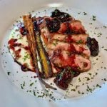 A gourmet plate featuring sliced duck breast with a rich berry sauce, accompanied by roasted carrots and a creamy white puree. Garnished with fresh herbs on a white plate.