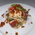 A plate of spaghetti topped with fig slices, shredded green herbs, and crispy bacon bits on a white plate. The dish is arranged neatly, showcasing the colorful ingredients on a plain table.