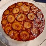 A round plum upside-down cake on a glass plate. The cake features a glossy caramelized top with evenly arranged plum slices, displaying a rich amber hue. It is set on a beige tablecloth under sunlight.