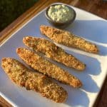 Four breaded and fried pickle spears on a square white plate, served with a small dish of creamy dipping sauce, are displayed on a wooden table in sunlight.