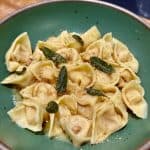 A green bowl filled with tortellini, topped with crispy sage and a sprinkle of cheese, placed on a wooden surface. The pasta is golden brown, glazed with a light sauce.
