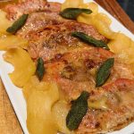 A rectangular white plate displaying a dish of four cooked meat slices, each topped with caramelized brown sauce. Slices of cooked apple and scattered sage leaves surround the meat. The plate is set on a wooden surface.