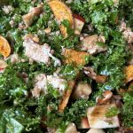 A bowl of kale salad with apple slices, roasted sweet potato wedges, walnuts, feta cheese, and sunflower seeds. The ingredients are tossed together, showcasing a mix of greens and vibrant colors.