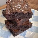 Three fudgy brownies stacked on a blue and white checkered plate, sprinkled with coarse sea salt. The brownies have a rich, dark chocolate appearance and a slightly crumbly texture on top.