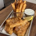 A serving tray with golden brown fish and chips, accompanied by a lemon wedge and a small cup of tartar sauce. The chips are held in a small metal basket, and everything is placed on a wooden table.