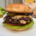 A juicy cheeseburger with two beef patties, melted cheese, pickles, tomato, and lettuce in a toasted bun. It sits on a white plate with a blurry background featuring lettuce and condiment bottles.