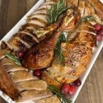 A platter of sliced roasted turkey with golden skin, garnished with fresh rosemary sprigs and scattered cranberries, is displayed on a wooden table. The turkey legs are whole and positioned in the center.