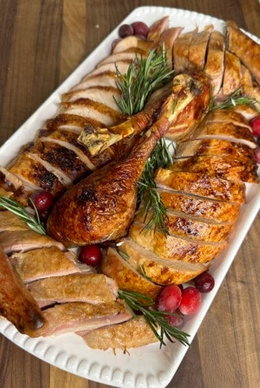 A platter of sliced roasted turkey with golden skin, garnished with fresh rosemary sprigs and scattered cranberries, is displayed on a wooden table. The turkey legs are whole and positioned in the center.