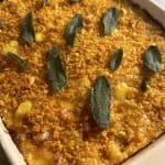 A close-up of a baked dish in a rectangular pan. The dish has a golden breadcrumb crust and is garnished with fresh sage leaves. The edges are slightly browned, suggesting its freshly baked.