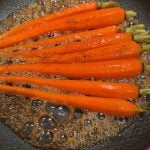 Bright orange carrots being sautéed in a frying pan with bubbling brown sauce. The carrots are evenly spaced, and the sauce appears to be well-seasoned with black pepper.