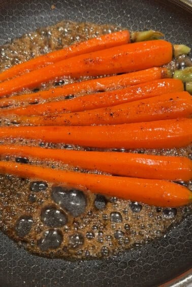 Bright orange carrots being sautéed in a frying pan with bubbling brown sauce. The carrots are evenly spaced, and the sauce appears to be well-seasoned with black pepper.