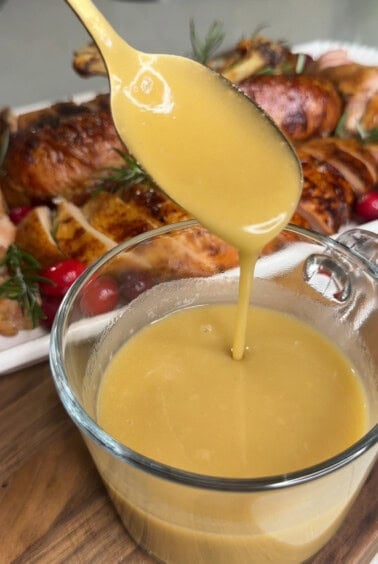 A golden spoon pours rich, creamy gravy into a glass container. In the background, roasted chicken garnished with fresh herbs and cranberries is visible, laid out on a white platter.