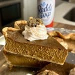 Close-up of a slice of pumpkin pie with whipped cream and a cookie crumble topping. Behind it is a can of Reddi-wip whipped cream, placed on a kitchen counter. Another pie slice is partially visible in the foreground.