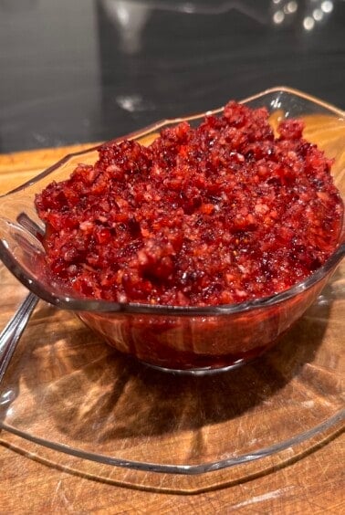 A glass dish filled with finely chopped cranberry relish, placed on a matching glass plate. A silver spoon is resting on the wooden surface next to it. The relish has a rich red color with a slightly coarse texture.