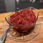 A glass bowl filled with a chunky, red cranberry relish sits on a matching glass plate atop a wooden surface. A metal spoon is placed beside the bowl.