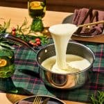 A saucepan filled with melted cheese is being lifted with a spoon, creating a long cheese pull. The table is set with a plaid tablecloth, brown napkins, and gold cutlery. Green glasses with drinks are decorated with orange slices.