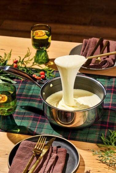 A saucepan filled with melted cheese is being lifted with a spoon, creating a long cheese pull. The table is set with a plaid tablecloth, brown napkins, and gold cutlery. Green glasses with drinks are decorated with orange slices.