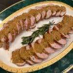 Sliced pork tenderloin drizzled with a brown sauce on a decorative green and gold-edged platter, garnished with rosemary sprigs.