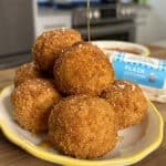 A plate stacked with golden-brown, deep-fried cheese balls, drizzled with honey. The cheese balls are crispy and garnished with a sprinkle of salt. A kitchen setting with blurred details is in the background.