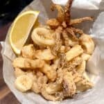 A cone-shaped paper holds deep-fried calamari with a wedge of lemon on the side. The calamari is golden brown and crispy, consisting of rings and tentacle pieces. The setup is on a wooden surface.