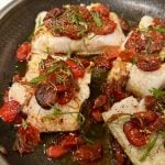 A close-up of a pan-seared fish filet topped with roasted cherry tomatoes and fresh herbs, all in a hot skillet. The dish appears well-cooked and garnished with finely chopped green herbs.