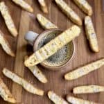 A cup of coffee on a wooden surface is surrounded by glazed biscotti. One biscotto rests across the cups rim. The biscotti are golden brown with a drizzle of white glaze.