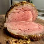 Close-up of a sliced roast beef on a wooden cutting board. The meat is cooked to medium-rare, showing a pink interior with a seasoned crust. A kitchen counter and some utensils are visible in the background.
