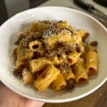 A white bowl filled with rigatoni pasta topped with crumbly bacon bits and grated cheese. The pasta is coated in a creamy sauce. The bowl is held in a hand above a wooden surface.