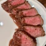 Sliced medium-rare steak with a brown sear, arranged on a white plate. The meat is juicy and topped with a light drizzle of sauce, showcasing a pink interior. The plate rests on a wooden table.