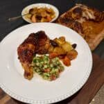 A plate with roasted chicken leg, diced cucumber and tomato salad, and roasted potatoes and carrots. In the background, a cutting board with more roasted chicken and a bowl with additional roasted vegetables.