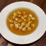 A white plate with a serving of tortellini in clear broth, set on a wooden table. The tortellini are small, pasta dumplings filled with ingredients, floating in the golden liquid.