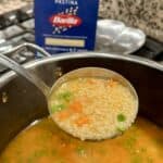 A ladle holds pastina soup over a pot. The soup contains small pasta, peas, and carrots in a clear broth. In the background, there is a box of Barilla Pastina on a kitchen counter.