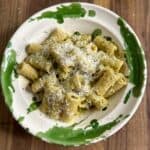 A plate of rigatoni pasta covered in pesto sauce and topped with grated cheese. The plate features a green splatter design around the rim and rests on a wooden table.